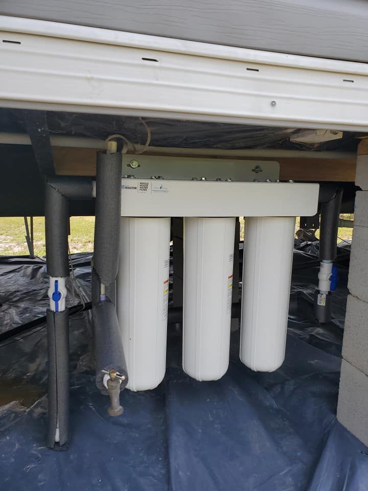 Three-stage water filter system installed under a structure, with black insulated pipes and a small spigot.