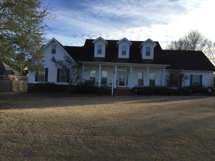 A two-story white house with a covered front porch and three roof dormers, set behind a dry, dormant grass lawn.