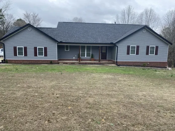 A single-story gray house with dark shutters, a brick base, and a front porch, set against a brown lawn and bare trees.