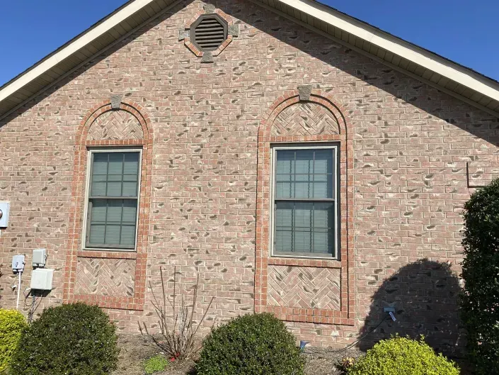 A brick house exterior with two arched windows, a circular vent, and green shrubs in front against a clear blue sky.