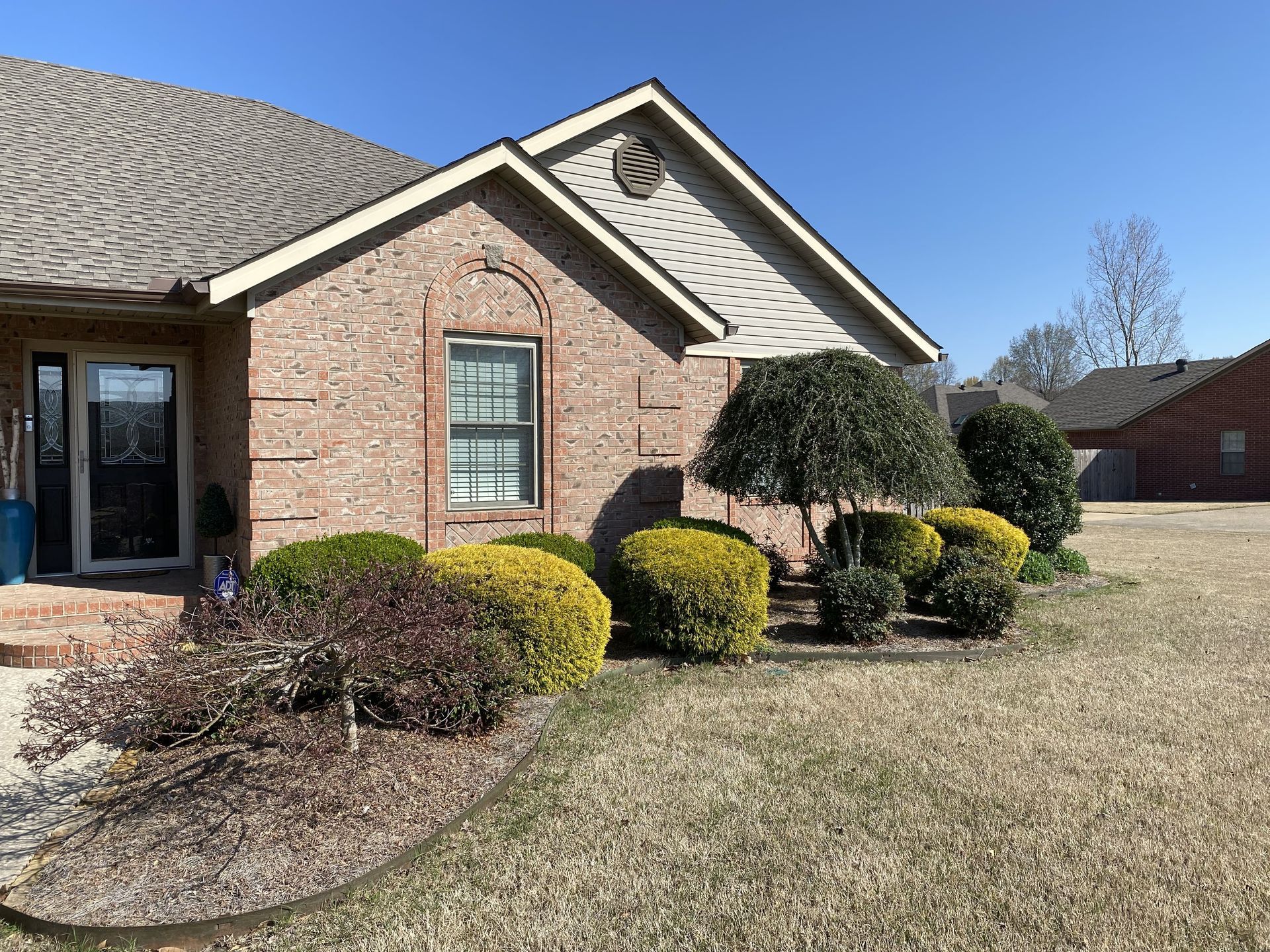 A brick house exterior with a front door, a side gable, and landscaped bushes under a clear blue sky.
