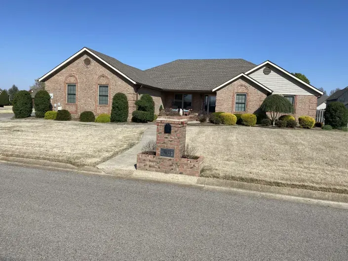 A tan brick, single-story suburban house with a brown roof and manicured bushes in the front yard under a clear blue sky.