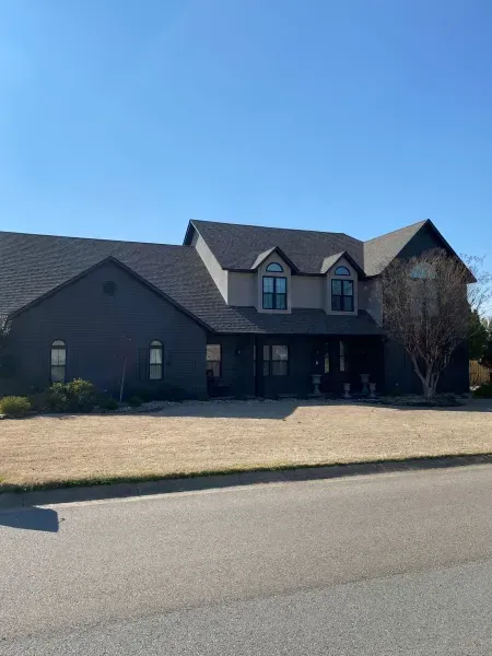 A two-story dark gray house with a peaked roof and two dormer windows sits behind a lawn under a clear blue sky.