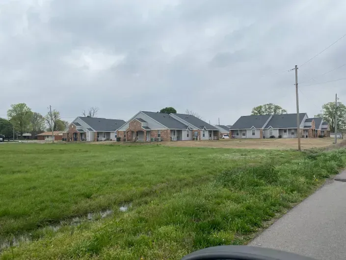 A row of newly constructed brick houses sits behind a large, grassy field under a cloudy sky.