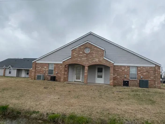 A brick-and-gray-siding duplex with two front doors under a shared arched entryway, set on a grassy lot under cloudy skies.