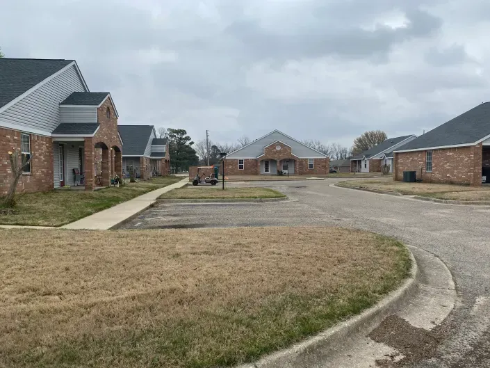 A row of one-story brick buildings with grey roofs lining a paved parking area and sidewalk under a cloudy sky.