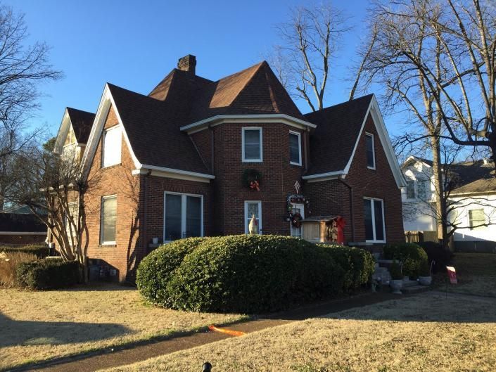A brick house with a prominent central turret, multiple gables, and a large trimmed shrub in the front yard.
