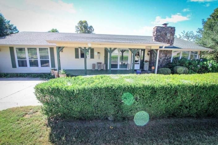 A cream-colored, single-story ranch house with a large stone chimney and a covered front porch behind a trimmed hedge.