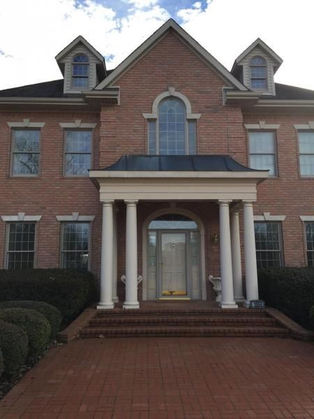 A two-story brick house with a front porch, white columns, arched windows, and two dormers under a partially cloudy sky.