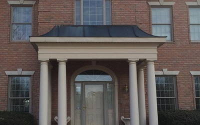 The front entrance of a two-story brick home featuring a dark metal porch roof supported by four white classical columns.