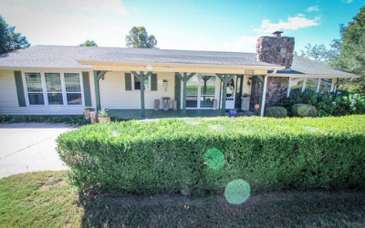 A one-story ranch house with cream siding, green shutters, a porch, and a large stone chimney set behind a manicured hedge.