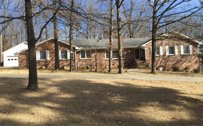 A one-story brick house with a gray roof and white shutters, situated on a grassy lot with many bare trees.