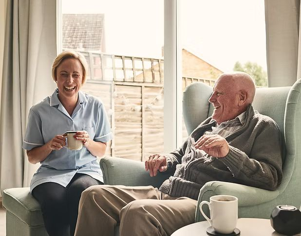 Two people sit chatting in a bright living room, each holding a mug, with a tea cup on the table.