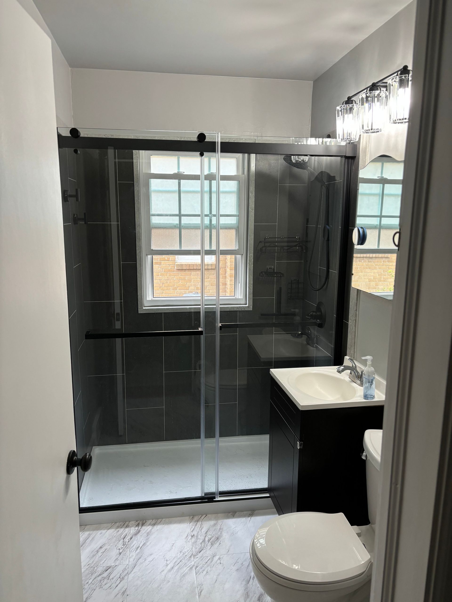 Bathroom with black tile shower, glass doors, window, dark vanity, and white toilet.
