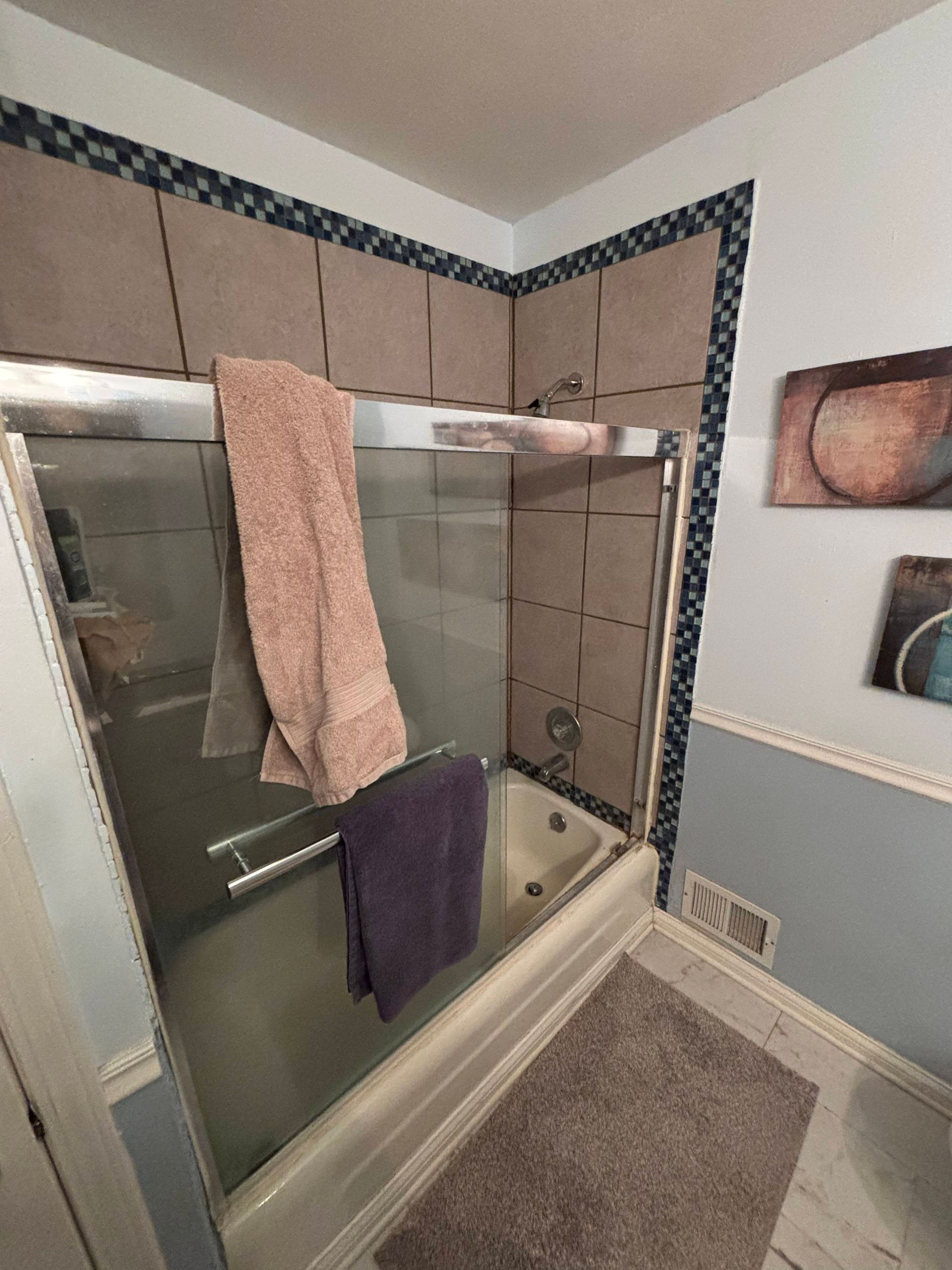 Shower with glass door, beige tile, blue trim, and two towels hanging inside.