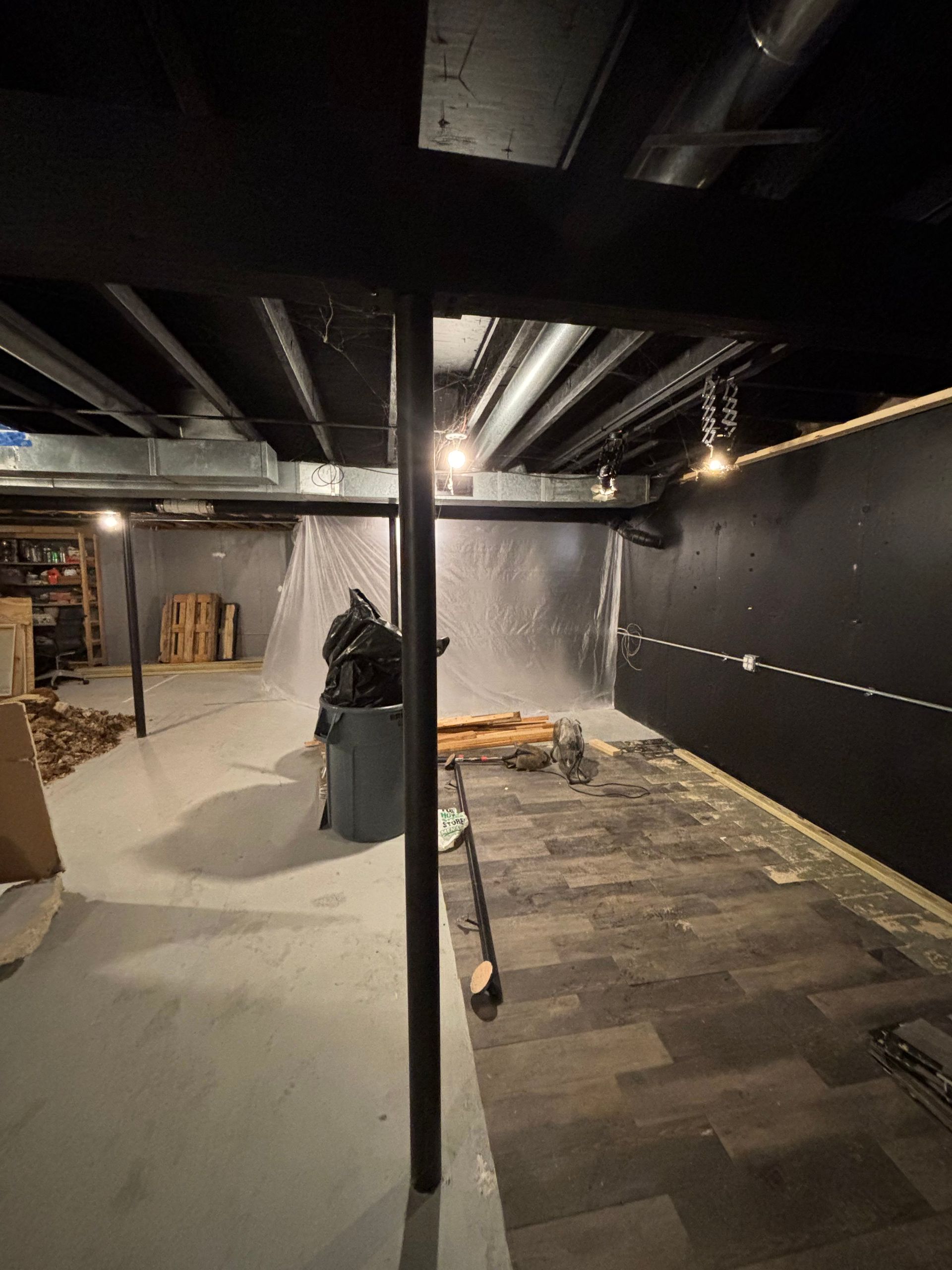 Basement renovation in progress. Black support beams, pipes on ceiling, unfinished flooring, gray walls.