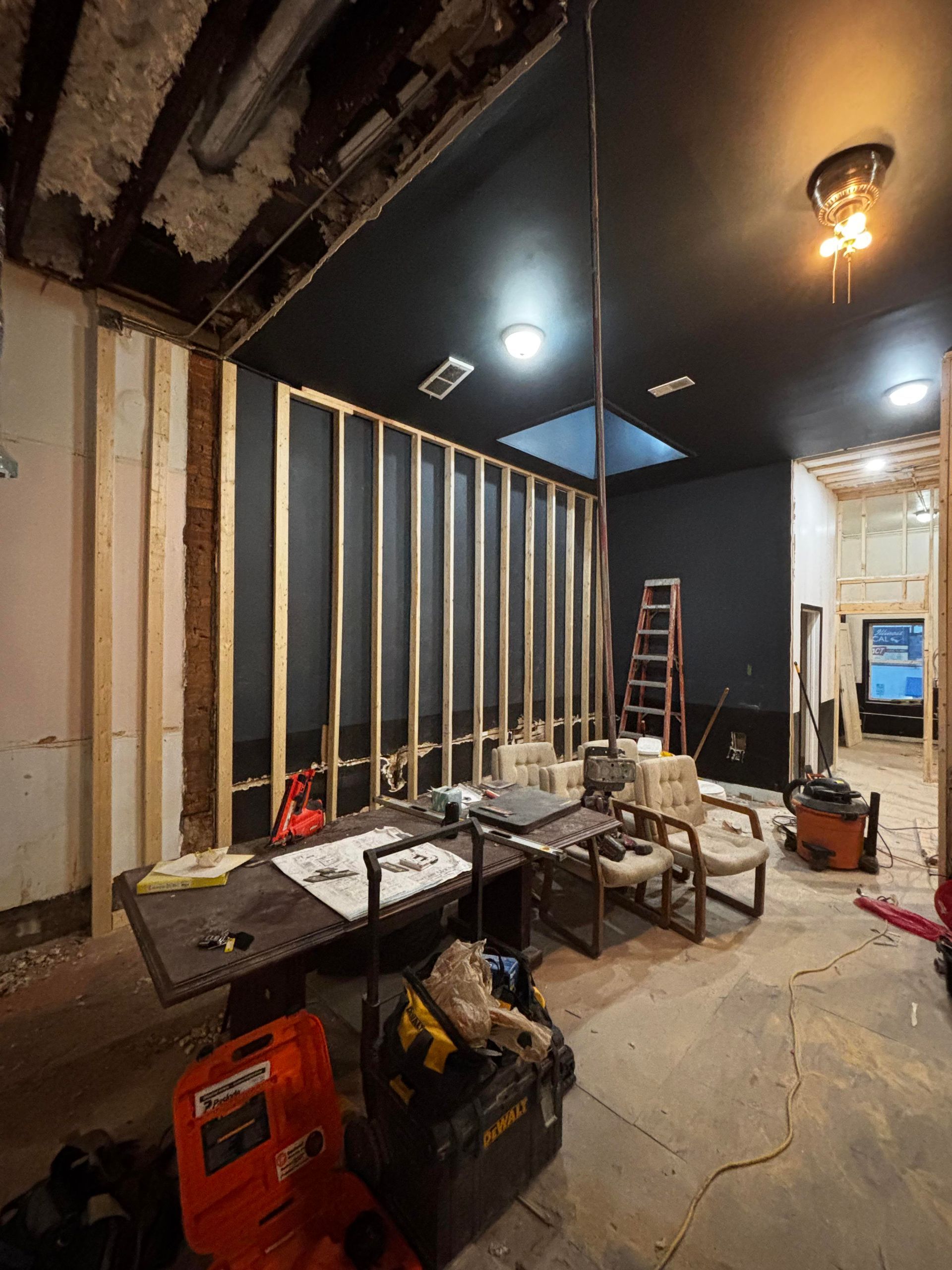 Interior undergoing renovation, exposed studs, black ceiling, desk with tools, chairs, ladder.