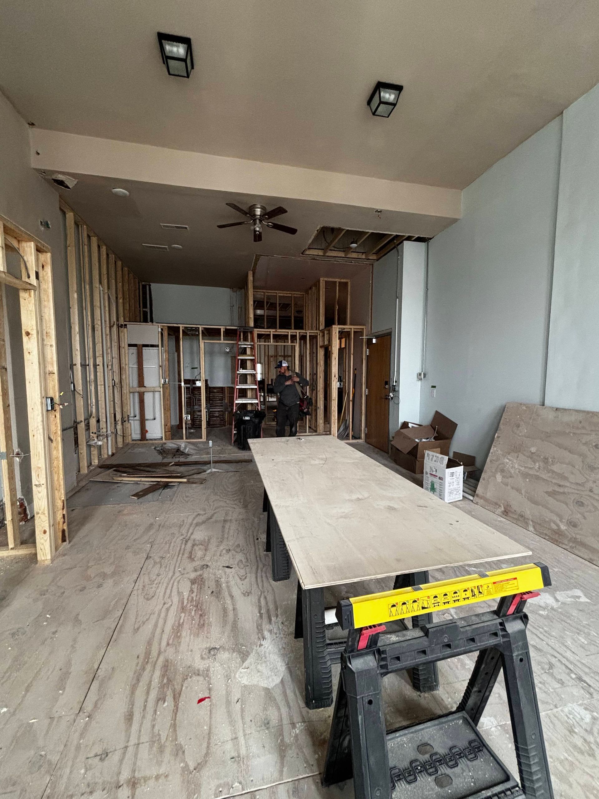 Interior construction site: framing, plywood on sawhorses, ceiling fixtures, and a person in the distance.