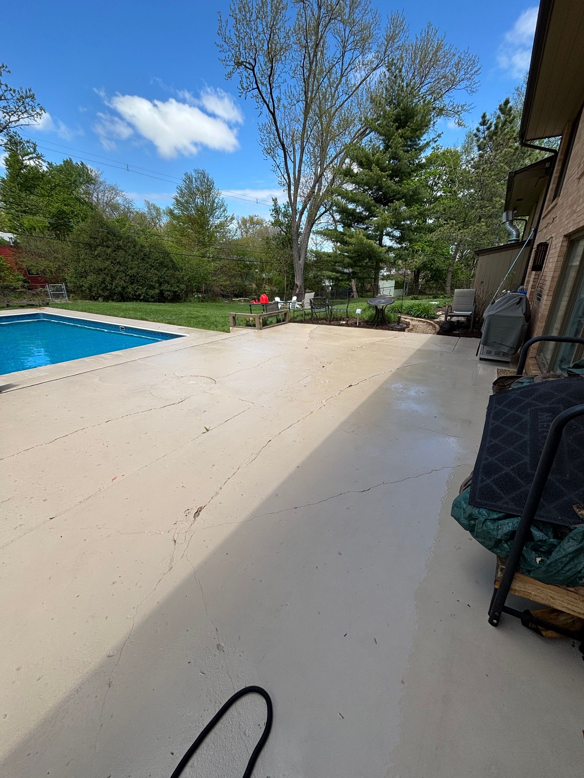 Concrete patio next to a pool and a house, with trees and a cloudy sky in the background.
