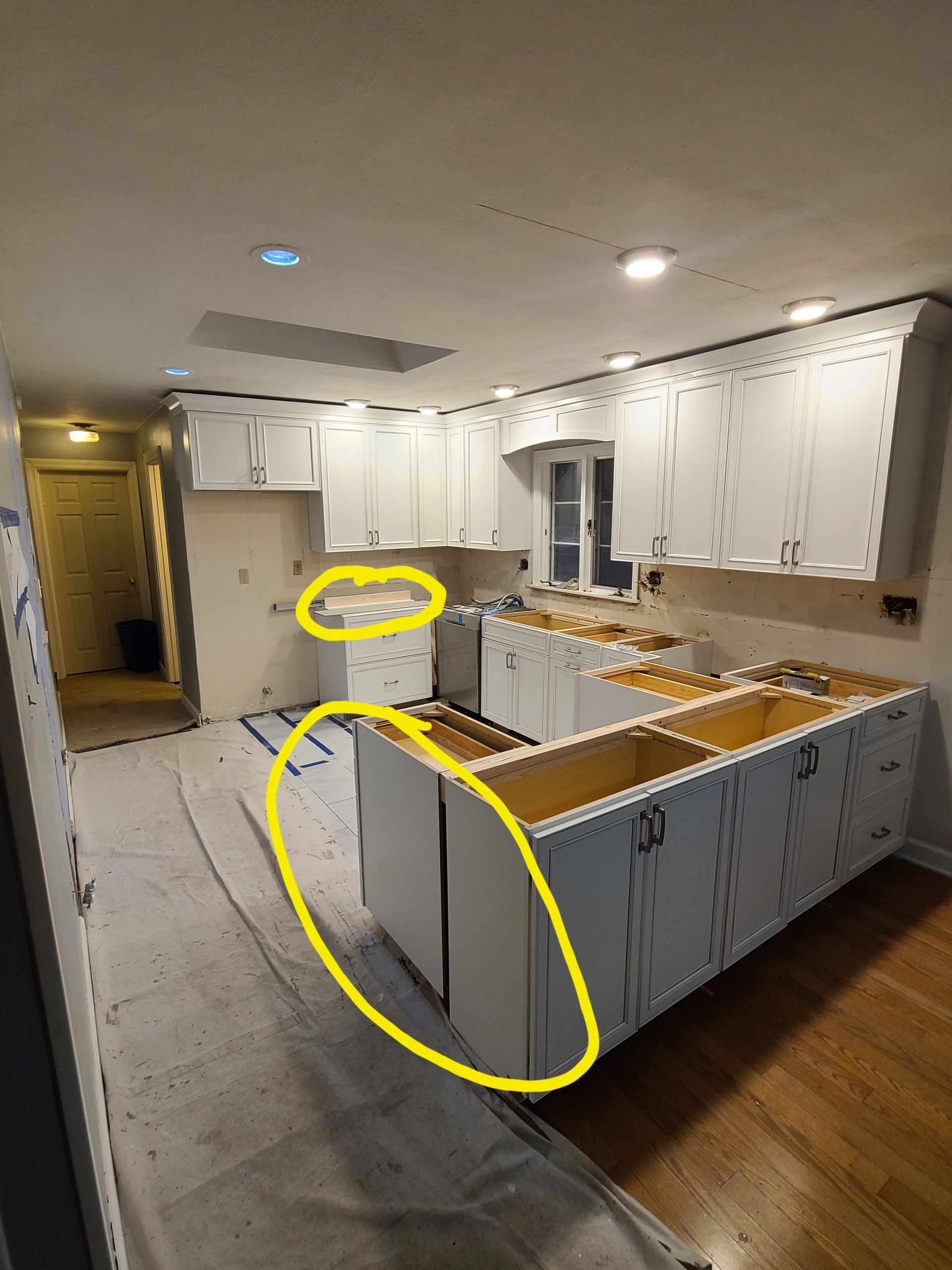 Kitchen cabinets under construction, white cabinets, island, and overhead lighting. Room covered in protective material.