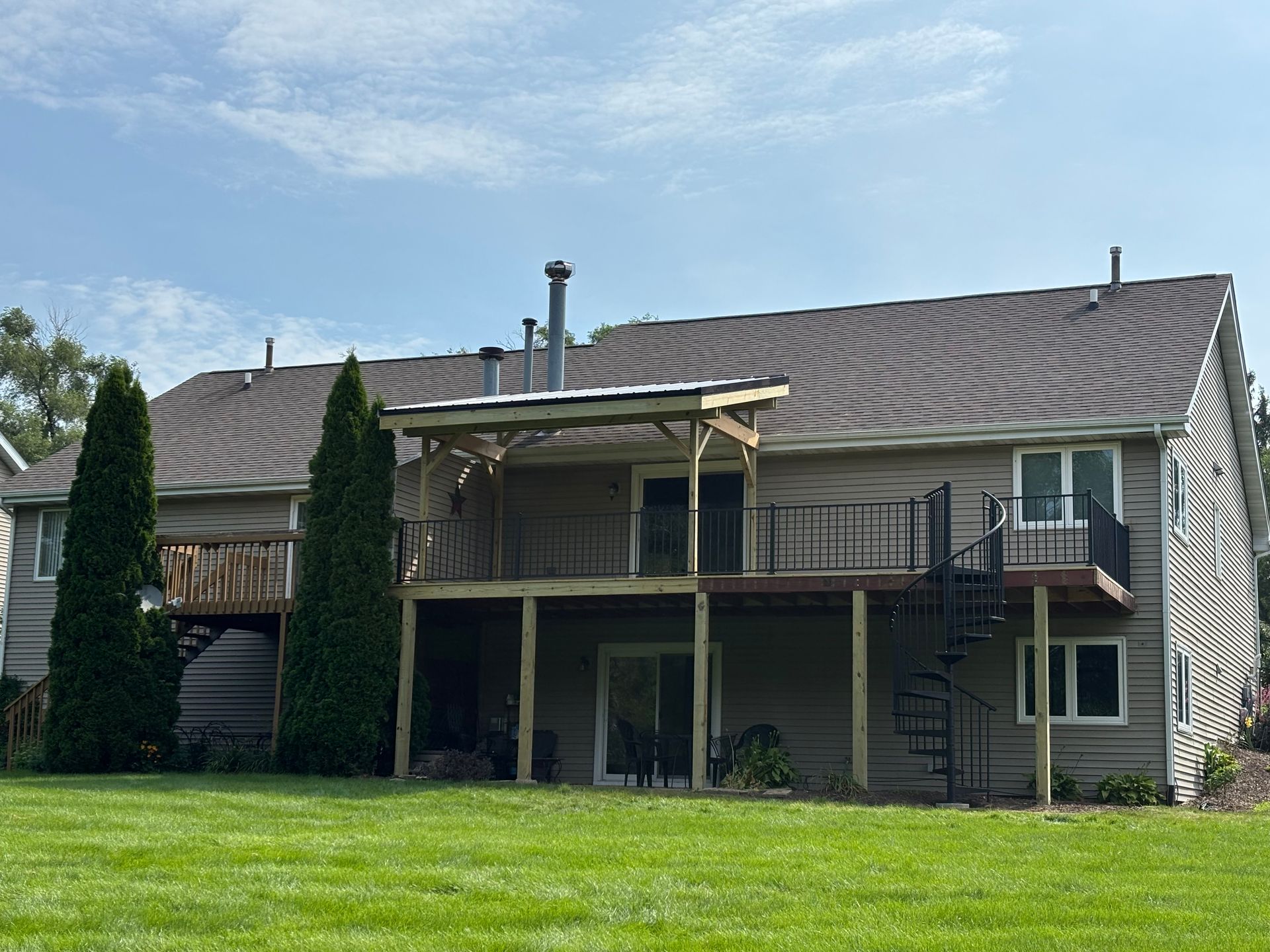 Back of a two-story house with a wooden deck, patio, and spiral staircase on a grassy lawn under a blue sky.