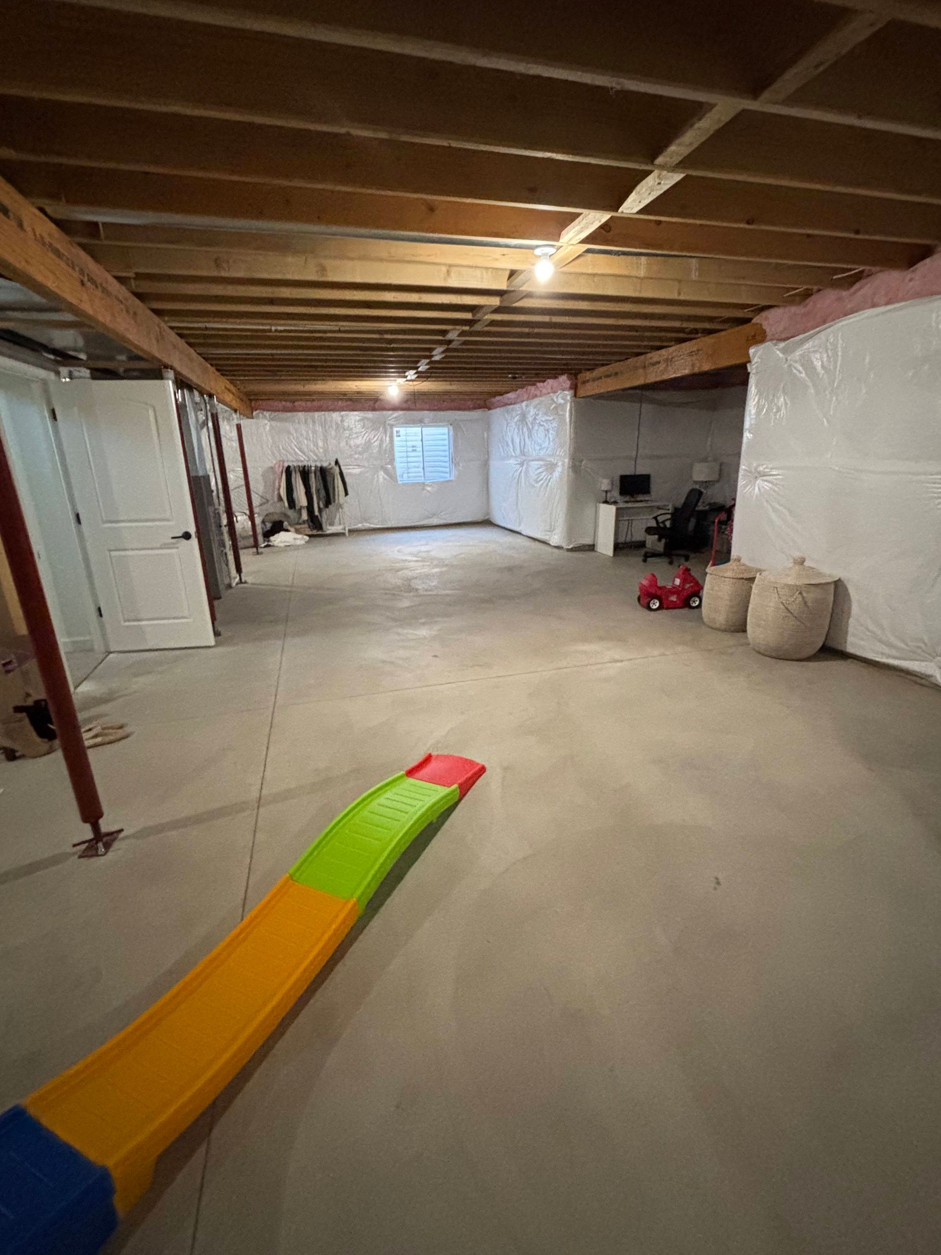 Basement interior. Concrete floor, exposed ceiling beams, plastic-covered walls. Children's slide in foreground.