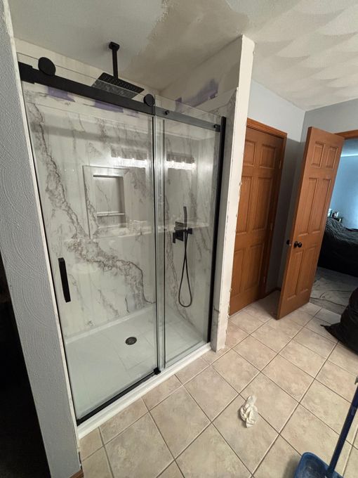 Shower with sliding glass doors, black hardware, marble-look wall. Beige tiled floor, wooden doors in background.