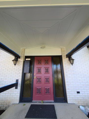 Red front door with decorative panels, flanked by sidelights. White brick exterior with a covered porch.