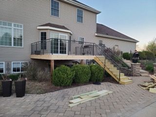 Backyard deck with black railings, stairs, and beige siding. Wooden planks lie on a brick patio.