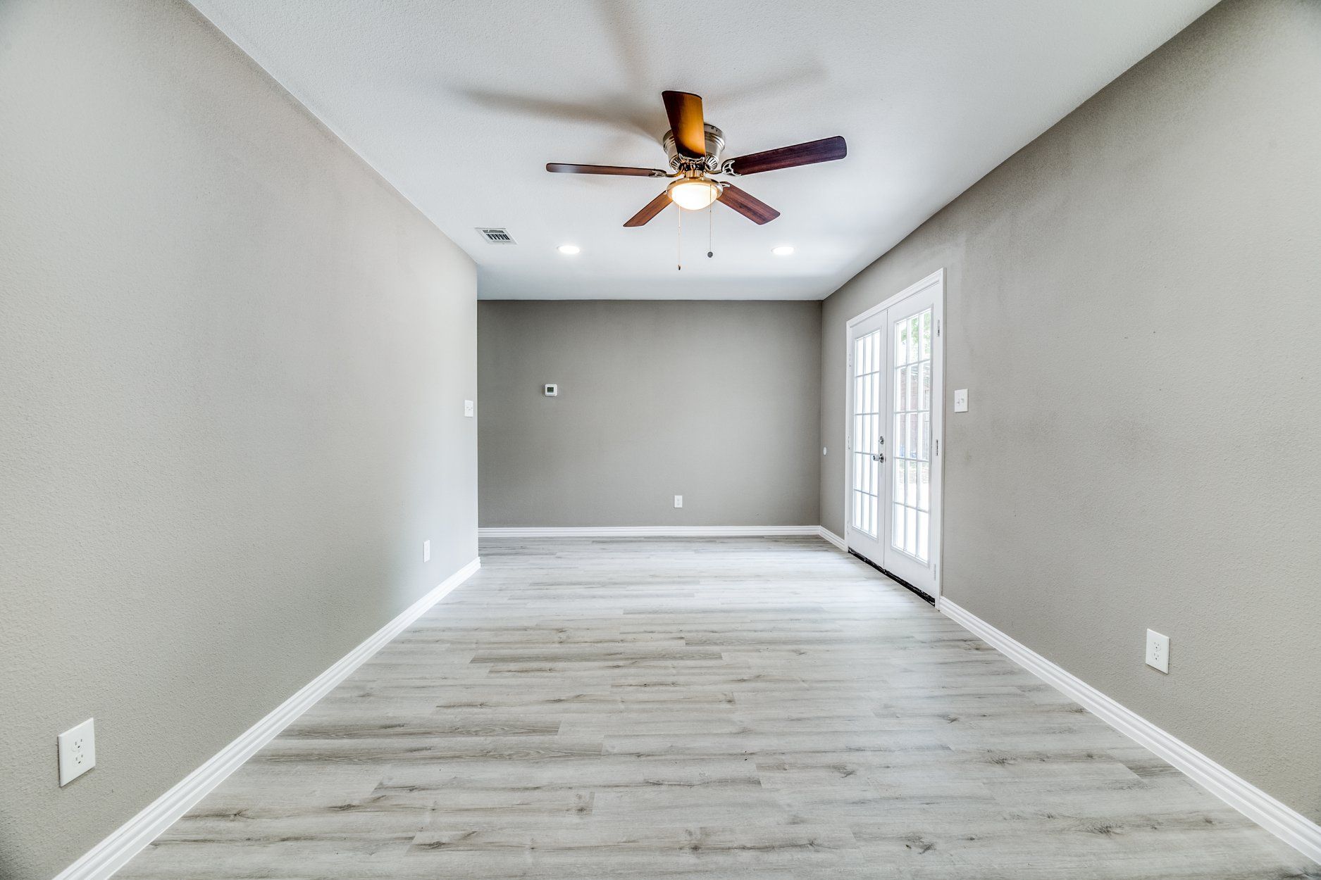 Empty room with gray walls, light wood-look floor, French doors, and ceiling fan.