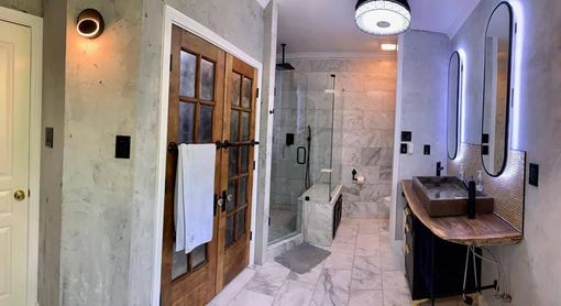 Bathroom with wooden doors, marble floor, shower, and two sinks with illuminated mirrors.