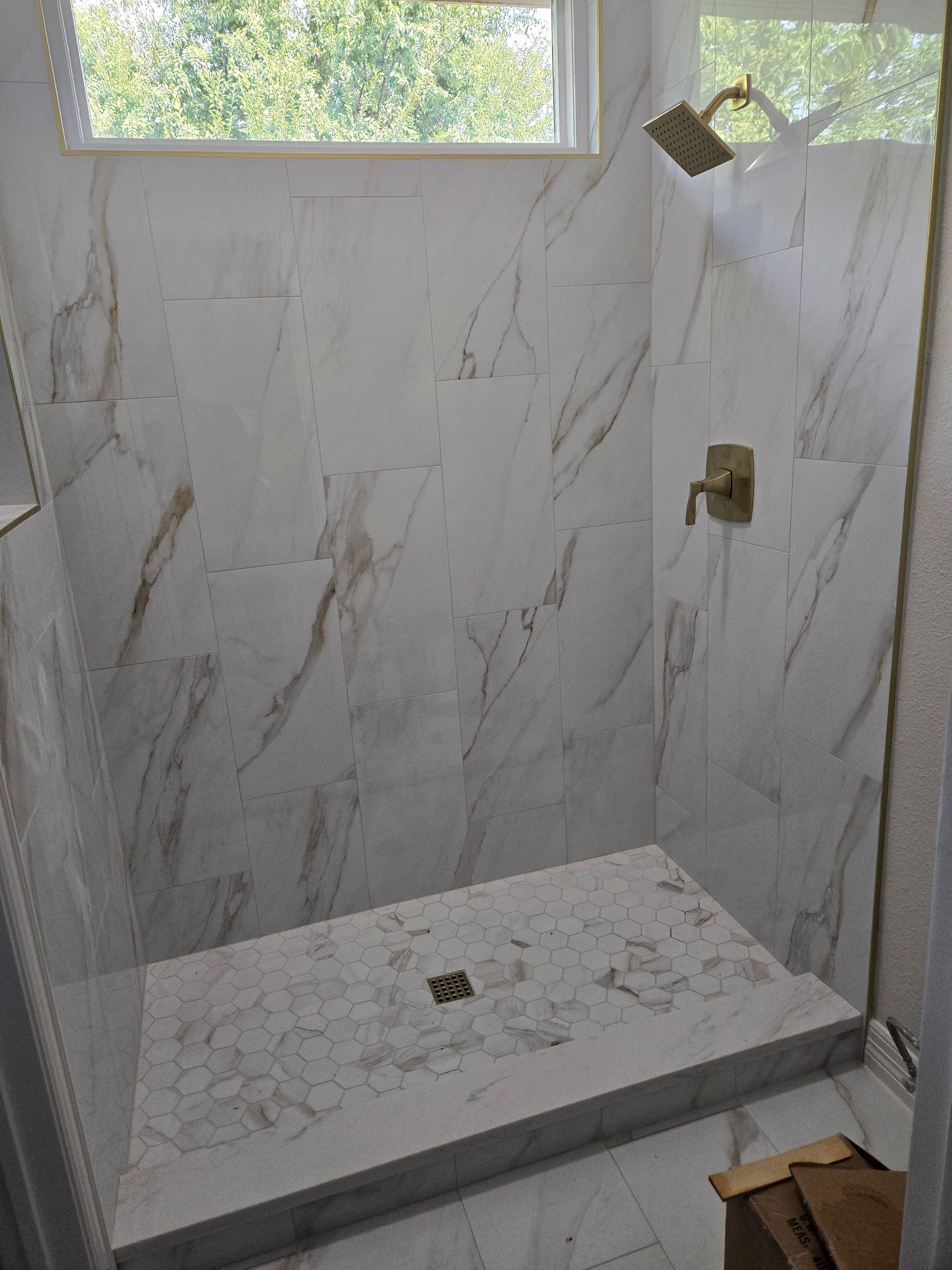 Shower with white marble-look tile, gold fixtures, and a glass door. Sunlight streams through a window.