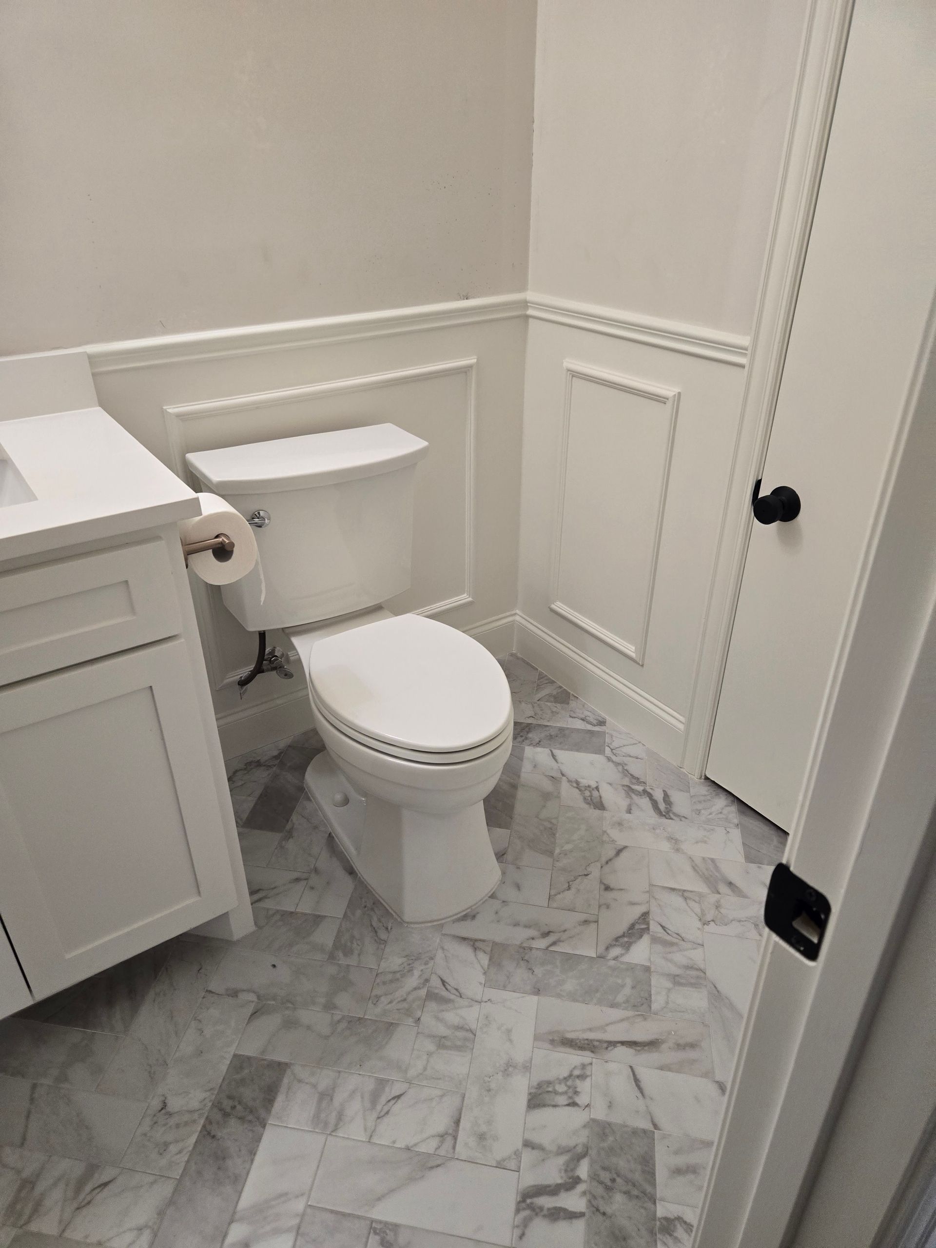 White bathroom with marble-patterned floor, toilet, vanity, and paneled walls.