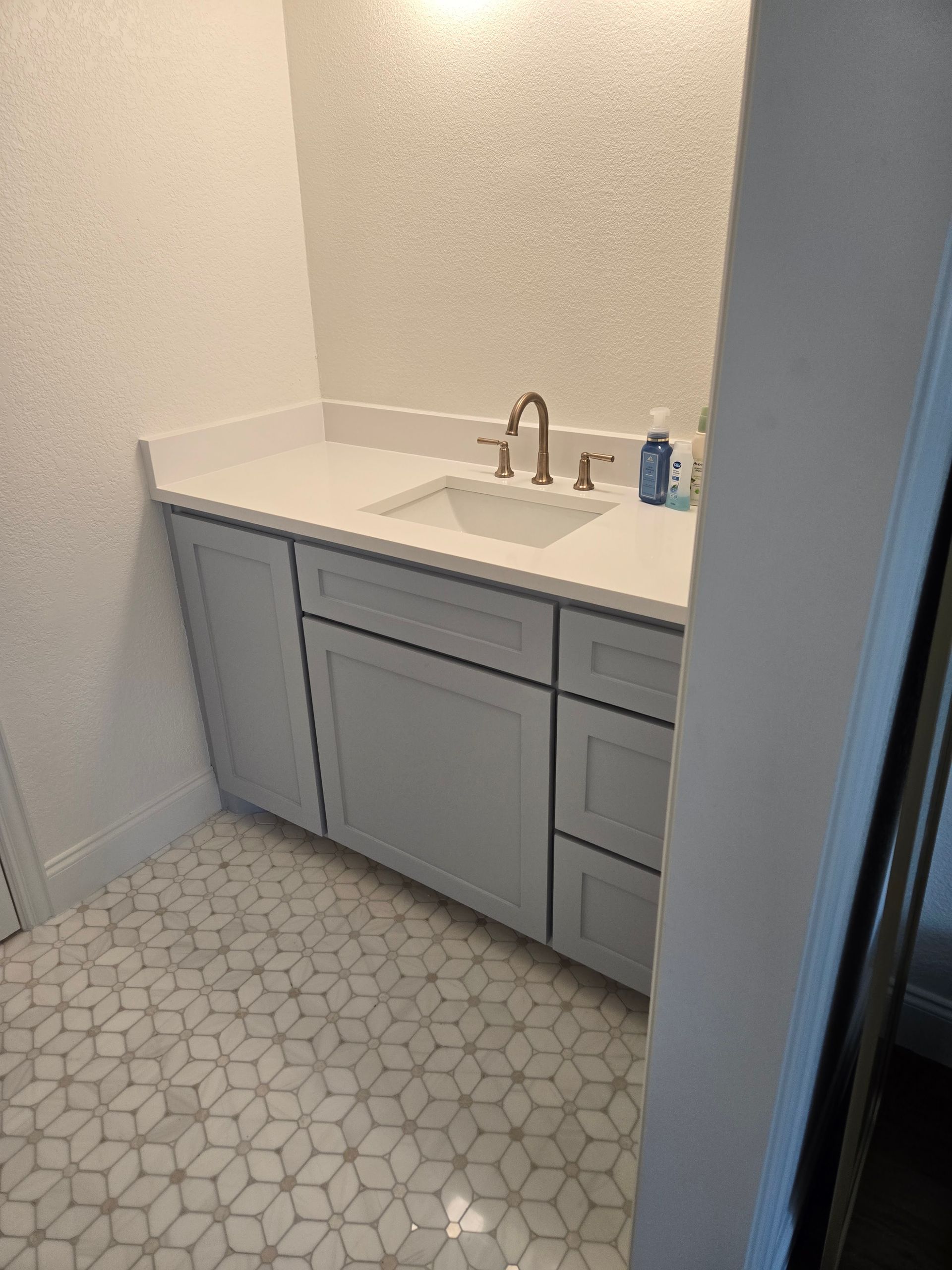 Bathroom with gray cabinets, white countertop, gold faucet, and patterned tile floor.