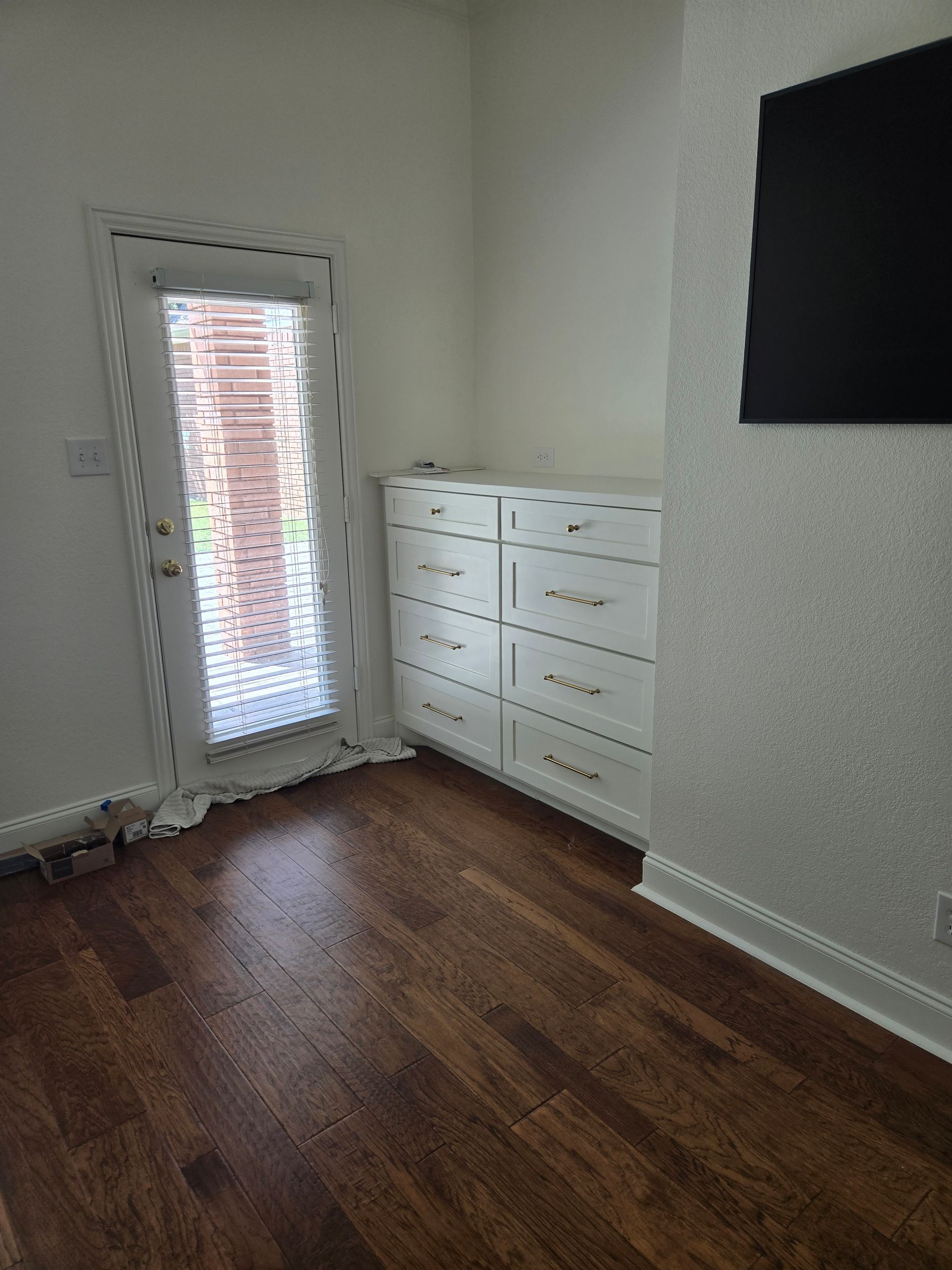Room with hardwood floor, white dresser, and glass door to outside. A TV is on the wall.