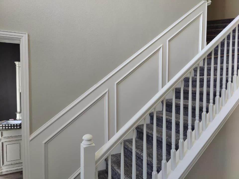 Staircase with white trim paneling and railing, gray carpeted stairs, and a doorway on the left.