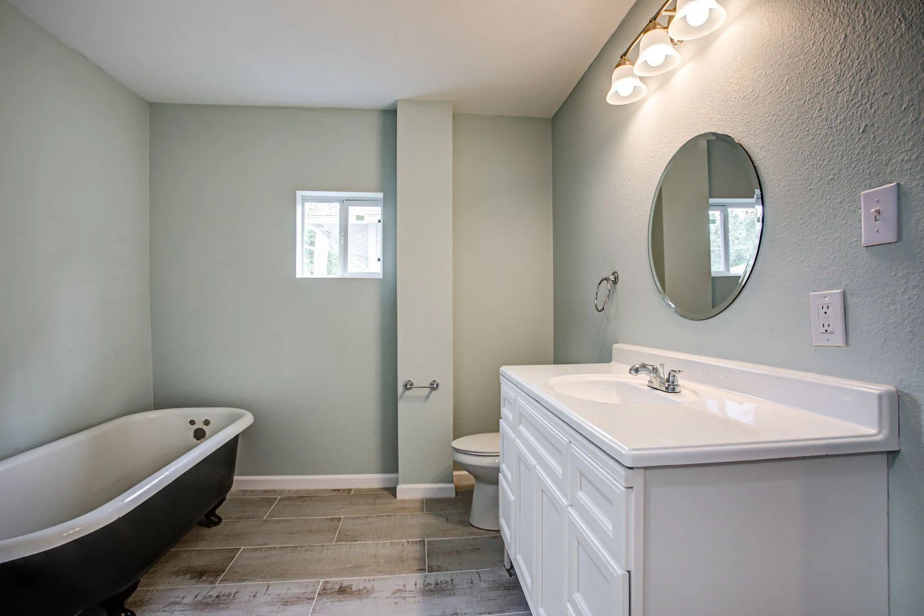 Bathroom with clawfoot tub, white vanity, round mirror, and pale green walls.