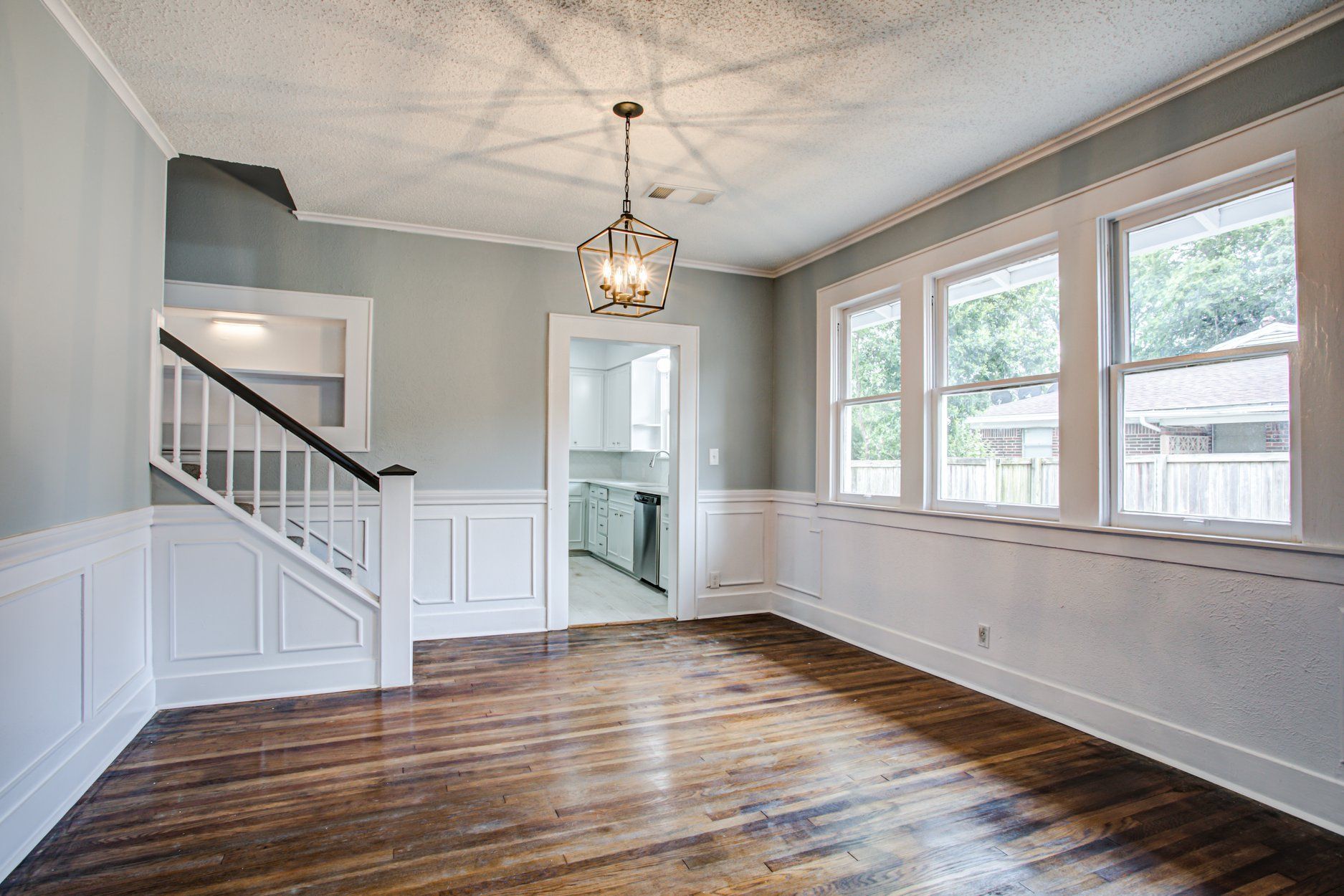 Empty room with hardwood floors, white trim, stairs, and windows. A chandelier hangs from the ceiling.