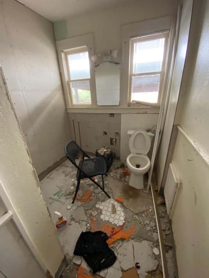 Dilapidated bathroom with damaged floor, toilet, two windows, and a chair.