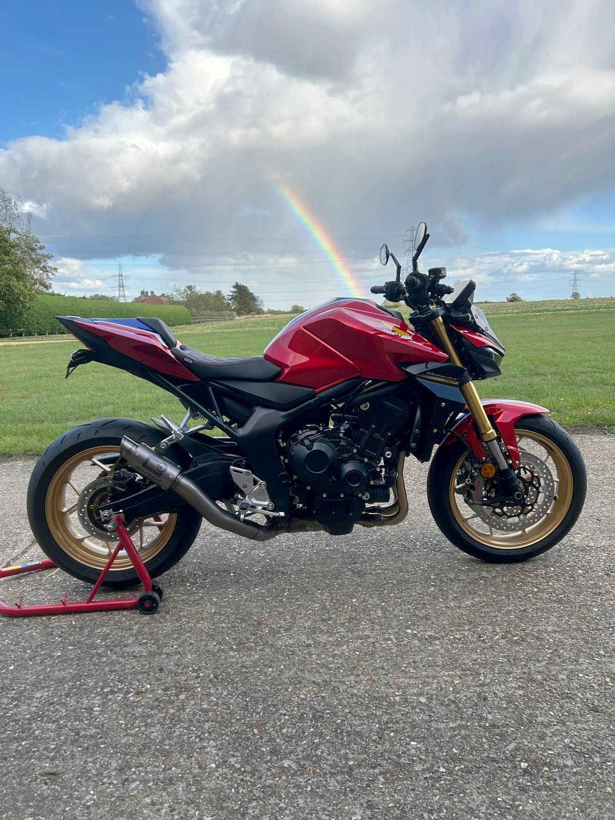 Red Honda motorcycle on a paved road, gold accents, rainbow in the background.