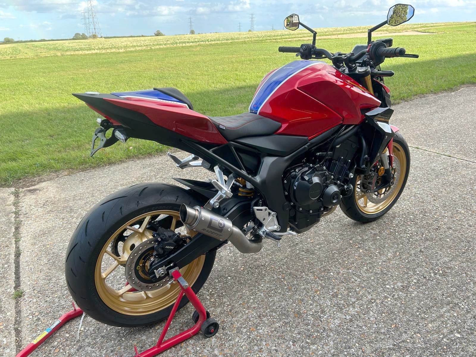 Red, blue, and black motorcycle with gold wheels on a stand, parked on pavement near grass.