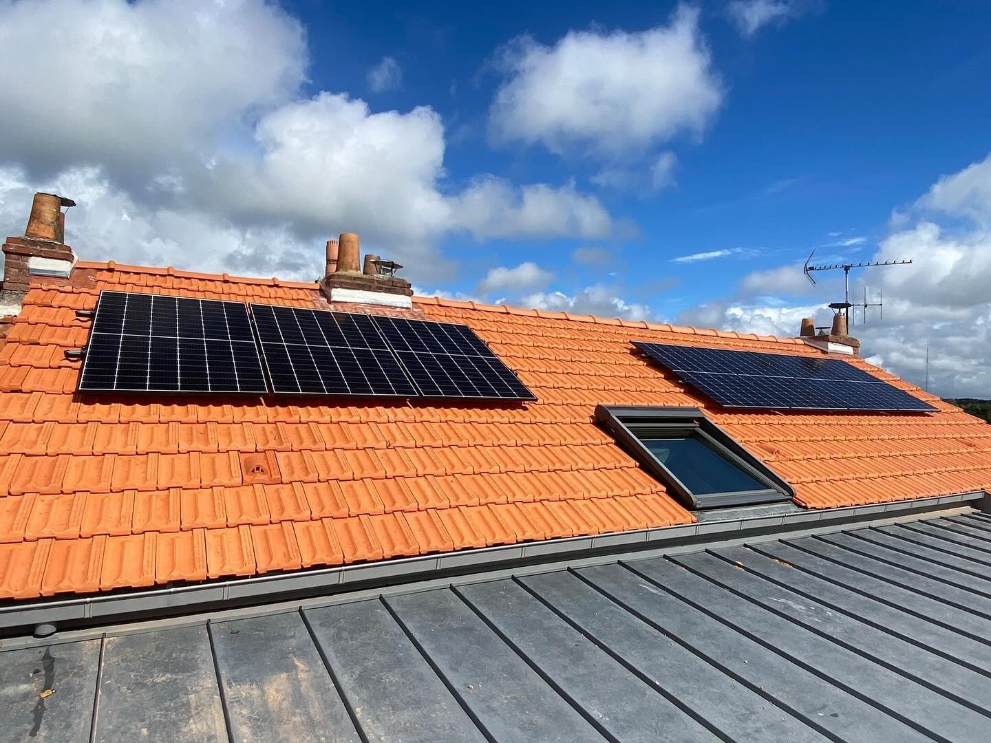 Toiture avec panneaux solaires noirs sur tuiles orange sous un ciel bleu nuageux