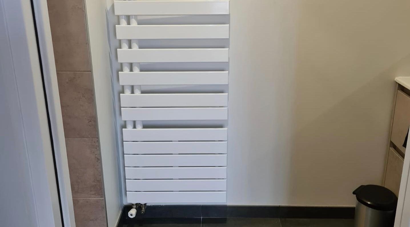 Cache-radiateur blanc à lattes à côté d'une porte dans une pièce beige, avec une petite poubelle sur la droite.