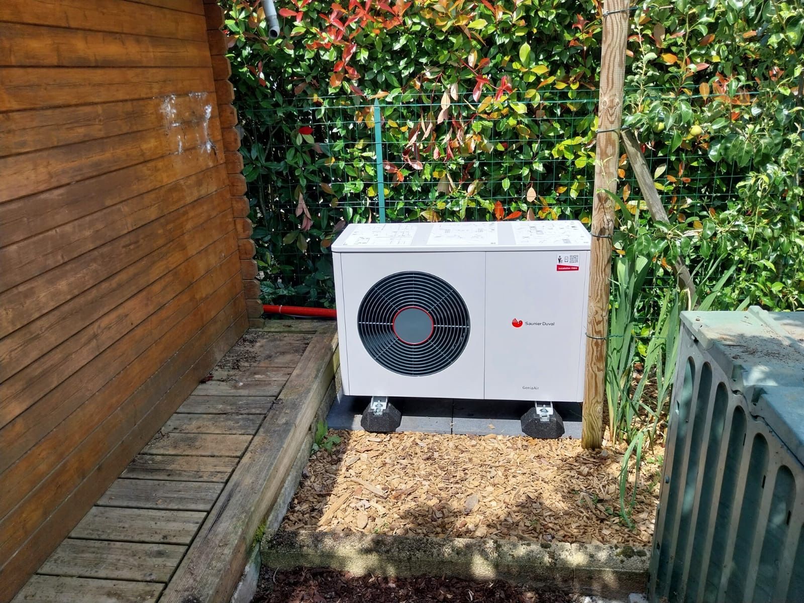 Unité de pompe à chaleur extérieure blanche, adossée à un mur en bois, installée sur une dalle de gravier dans un jardin.