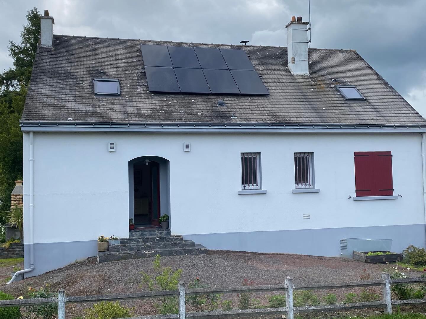 Maison blanche avec un toit gris, des panneaux solaires, un volet rouge et une porte ouverte dans un cadre rural