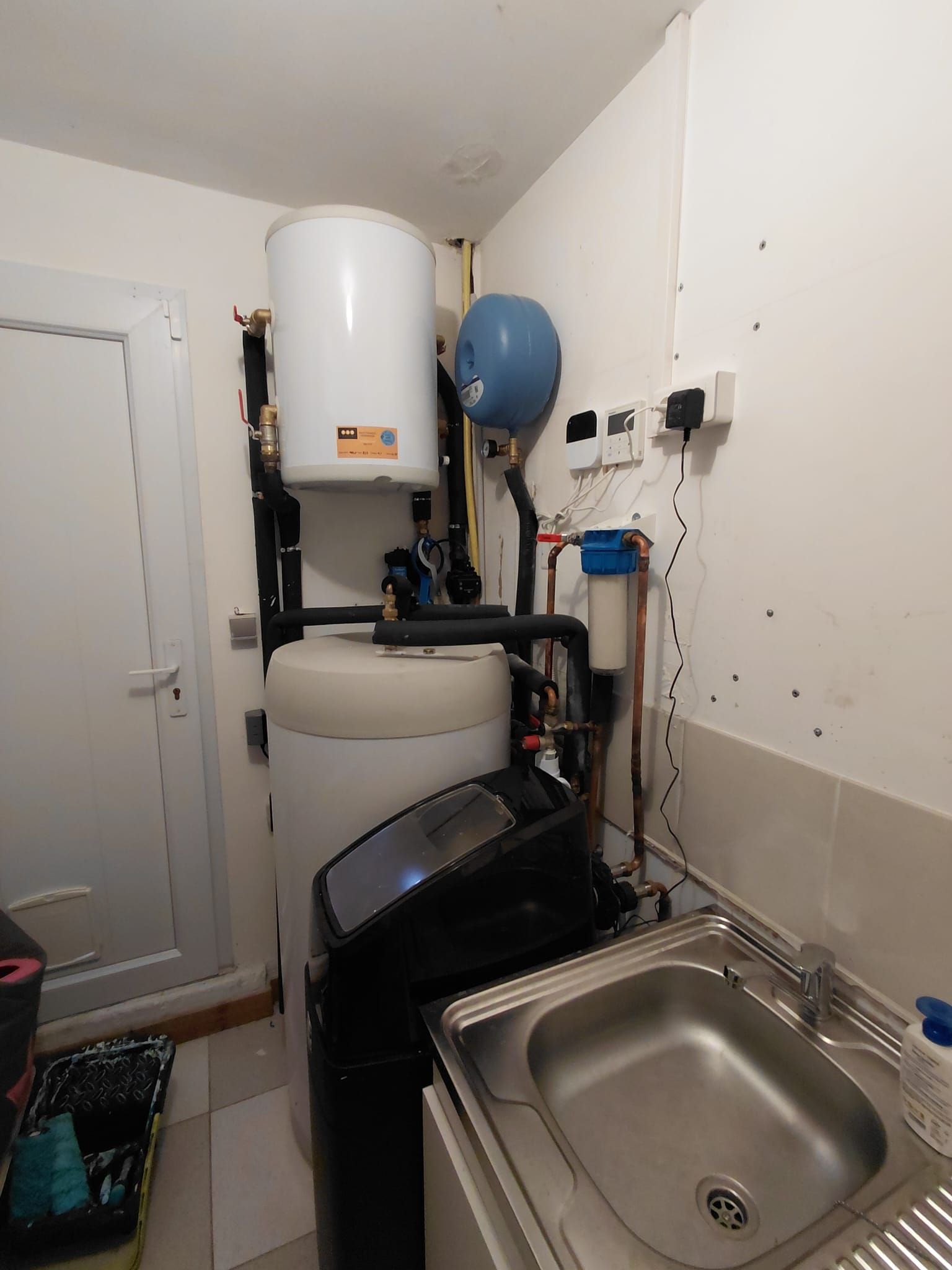 Petite buanderie avec réservoirs d'eau, canalisations, évier et matériel de nettoyage.