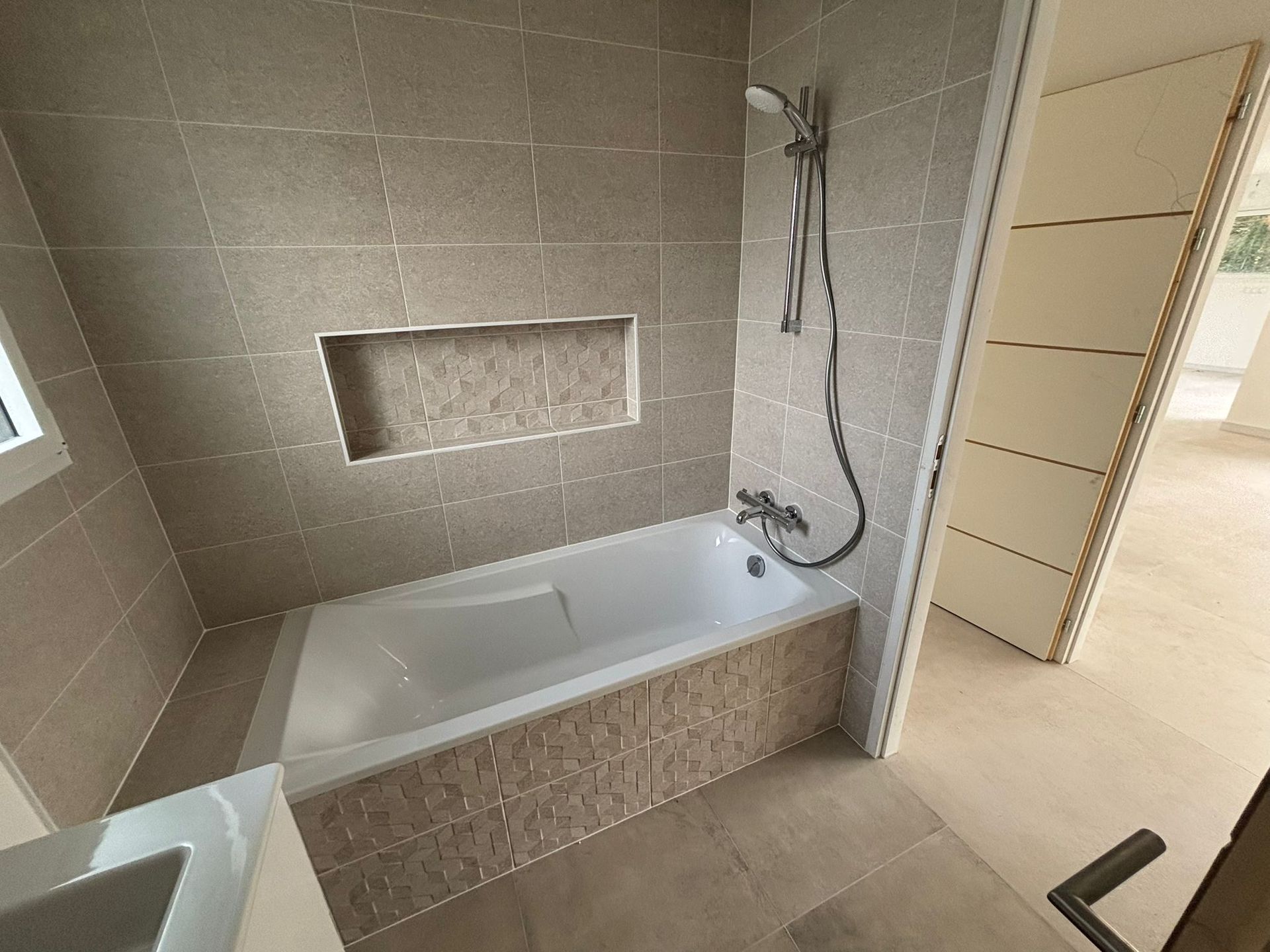 Salle de bains moderne avec baignoire, murs carrelés, douchette et niche d'étagère encastrée.