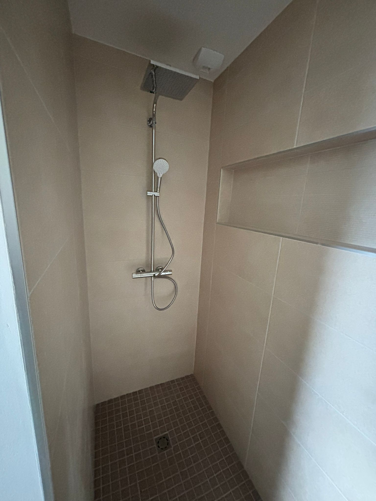 Petite cabine de douche beige avec douchette, barre de douche murale et sol carrelé.