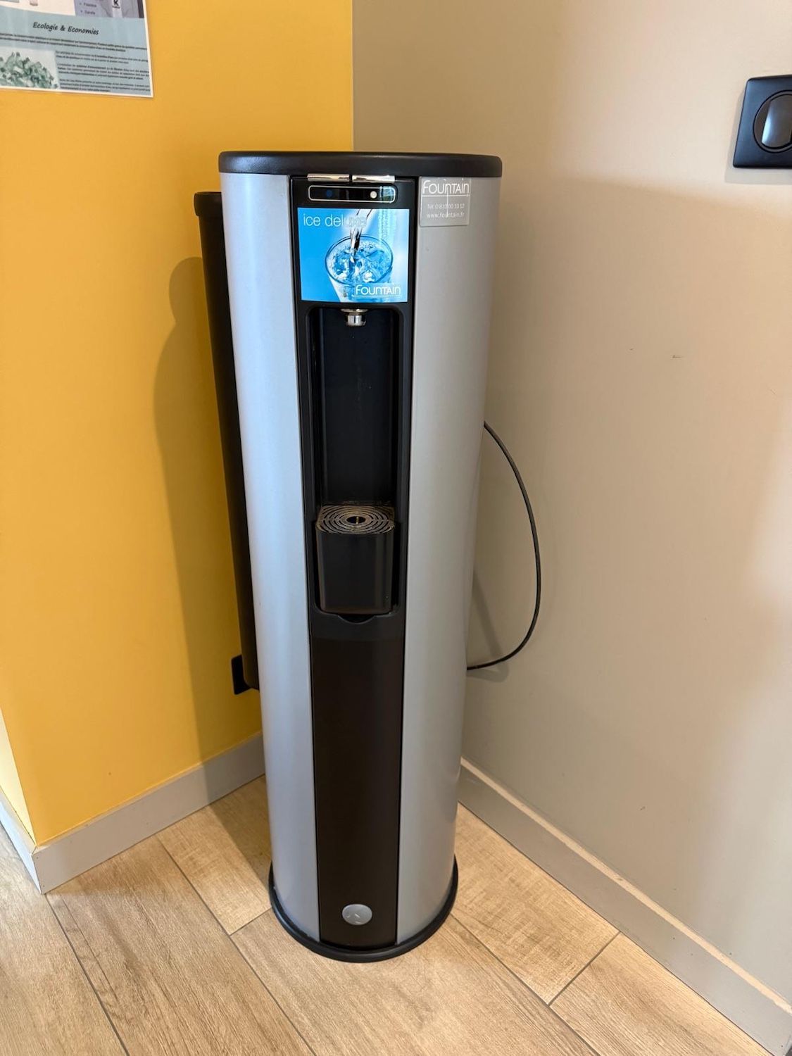 Fontaine à eau argentée dans un coin, contre des murs jaunes et beiges sur un parquet clair