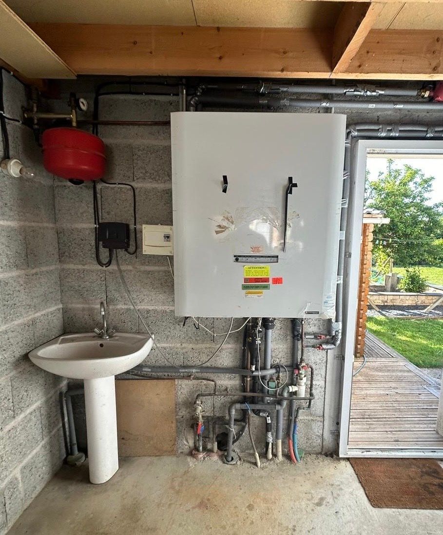 Buanderie avec chauffe-eau mural, évier, tuyauterie et porte ouverte sur l'extérieur.